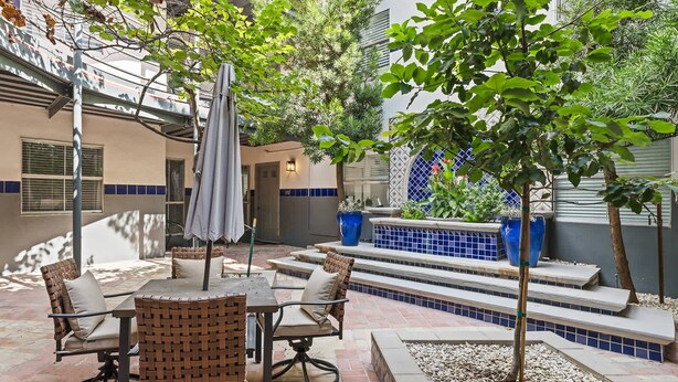 Une terrasse extérieure avec une table à manger, des chaises et un parasol. Des marches mènent à un espace détente agrémenté de carreaux bleus et de plantes en pot.