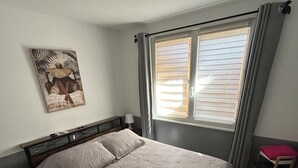 A bedroom with a bed, a nightstand, a window with blinds, a wall-mounted artwork, and a small shelf. - Gîte du Tacot 21 – A peaceful retreat in the heart of nature (Saulieu)