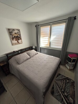 Ein Schlafzimmer mit einem Bett, einem Nachttisch, einem Fenster mit Jalousien, einem Wandbild und einer kleinen Ablage.