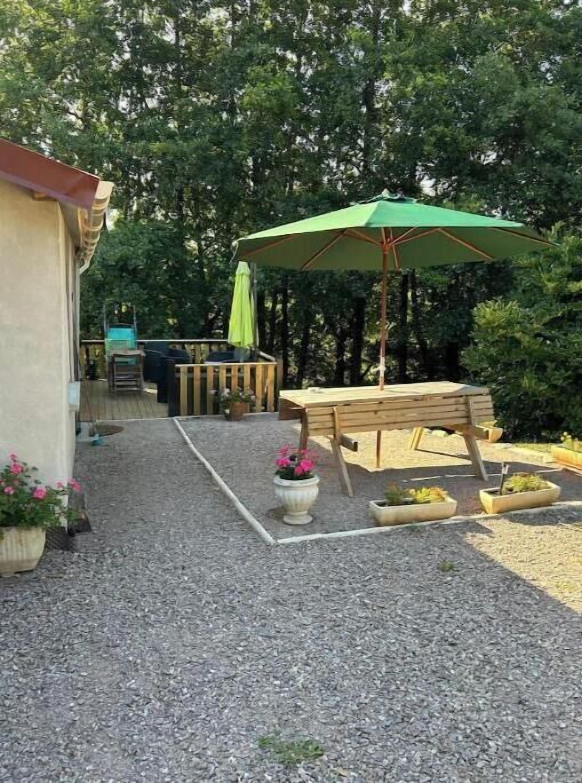 A patio area with a wooden table and bench, surrounded by gravel, potted plants, and a green umbrella.