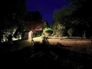 Ein beleuchtetes Areal im Freien bei Nacht mit einem Weg, Bäumen und einer kleinen, beleuchteten Struktur.