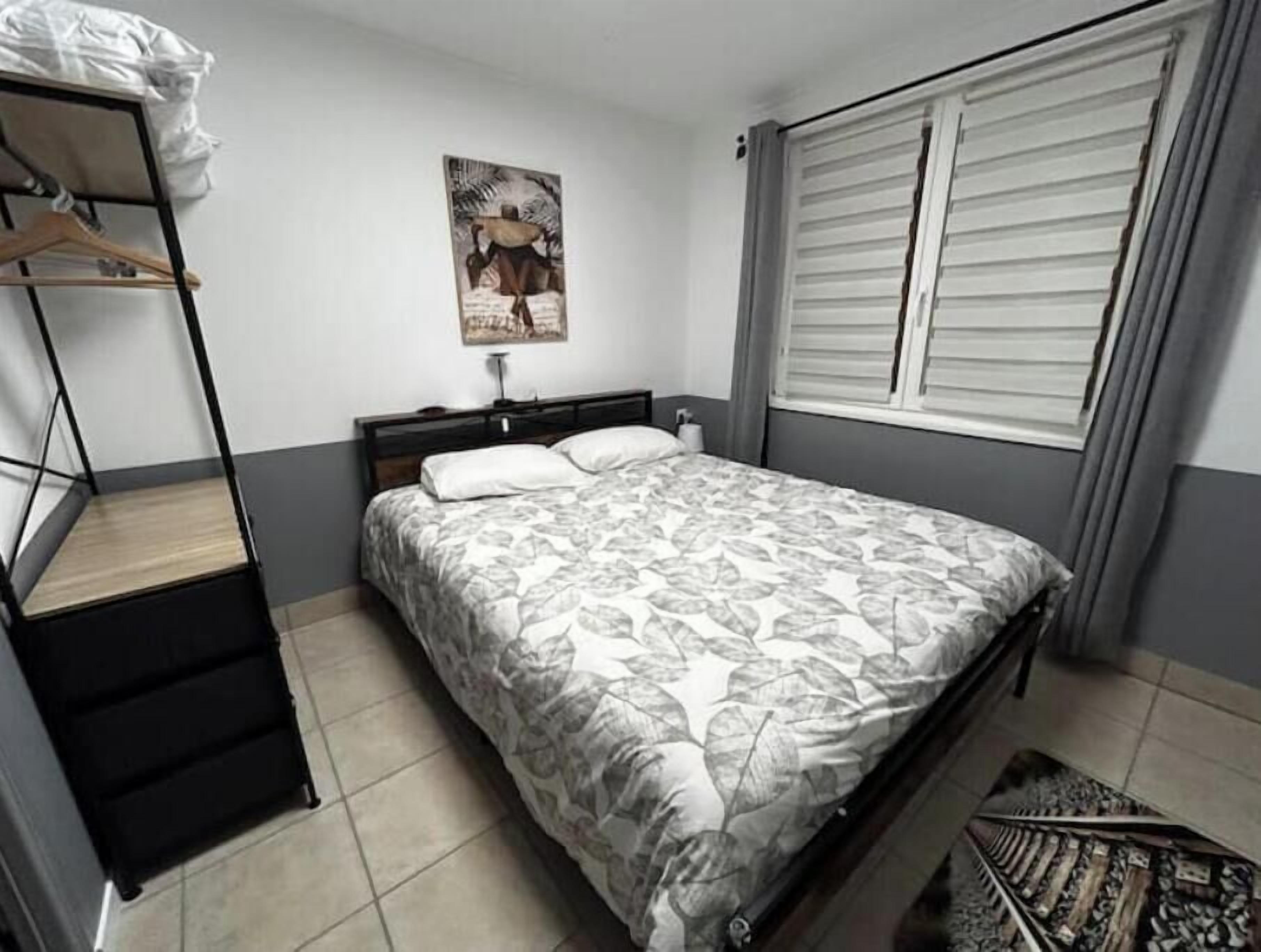 Ein Schlafzimmer mit Bett, Nachttisch, Wandbild, Fenster mit Jalousien und einem Etagenbett mit angehängter Tasche.