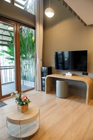 A modern living room with a TV, a coffee machine, a round table with flowers, and a view of greenery outside. - Cozy Mezzanine Loft Bedroom and Master Suite with Shared Pool & Co-Work Deck (badung)