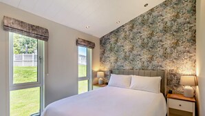 Un dormitorio con una cama grande, dos mesitas de noche con lámparas, papel tapiz floral y vistas a un jardín césped a través de una ventana.
