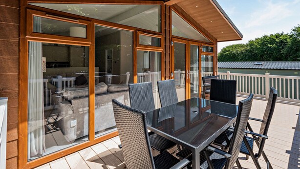 Un área de comedor exterior moderna con paredes de vidrio, mobiliario de mimbre y vistas al interior.