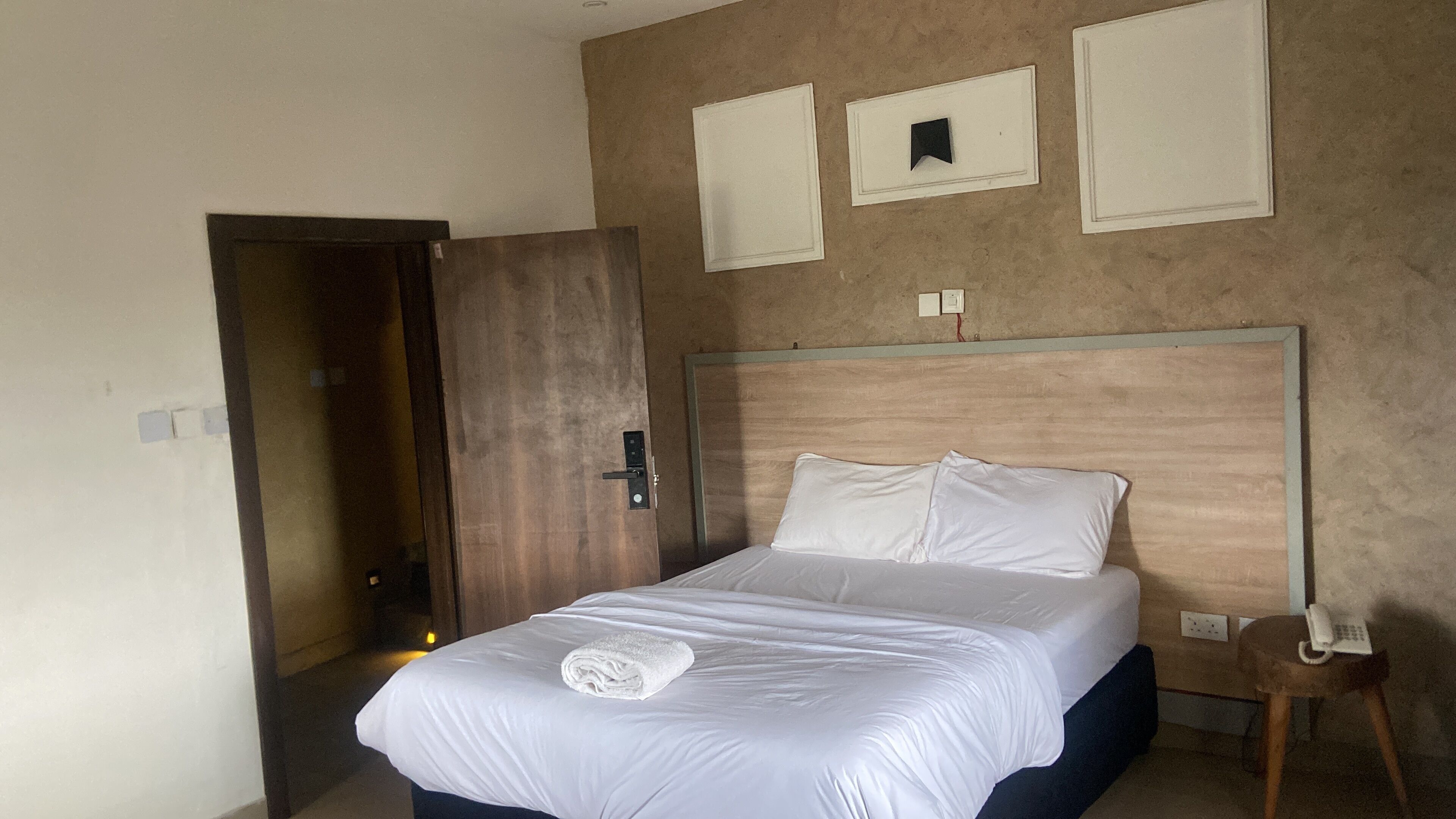 Quarto de hotel com uma cama, porta de madeira, mesa de cabeceira com telefone e interruptor de luz na parede.