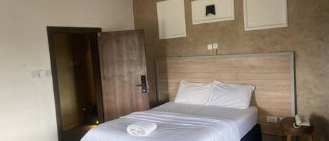 A hotel room with a bed, a wooden door, a bedside table with a phone, and a wall-mounted light switch.