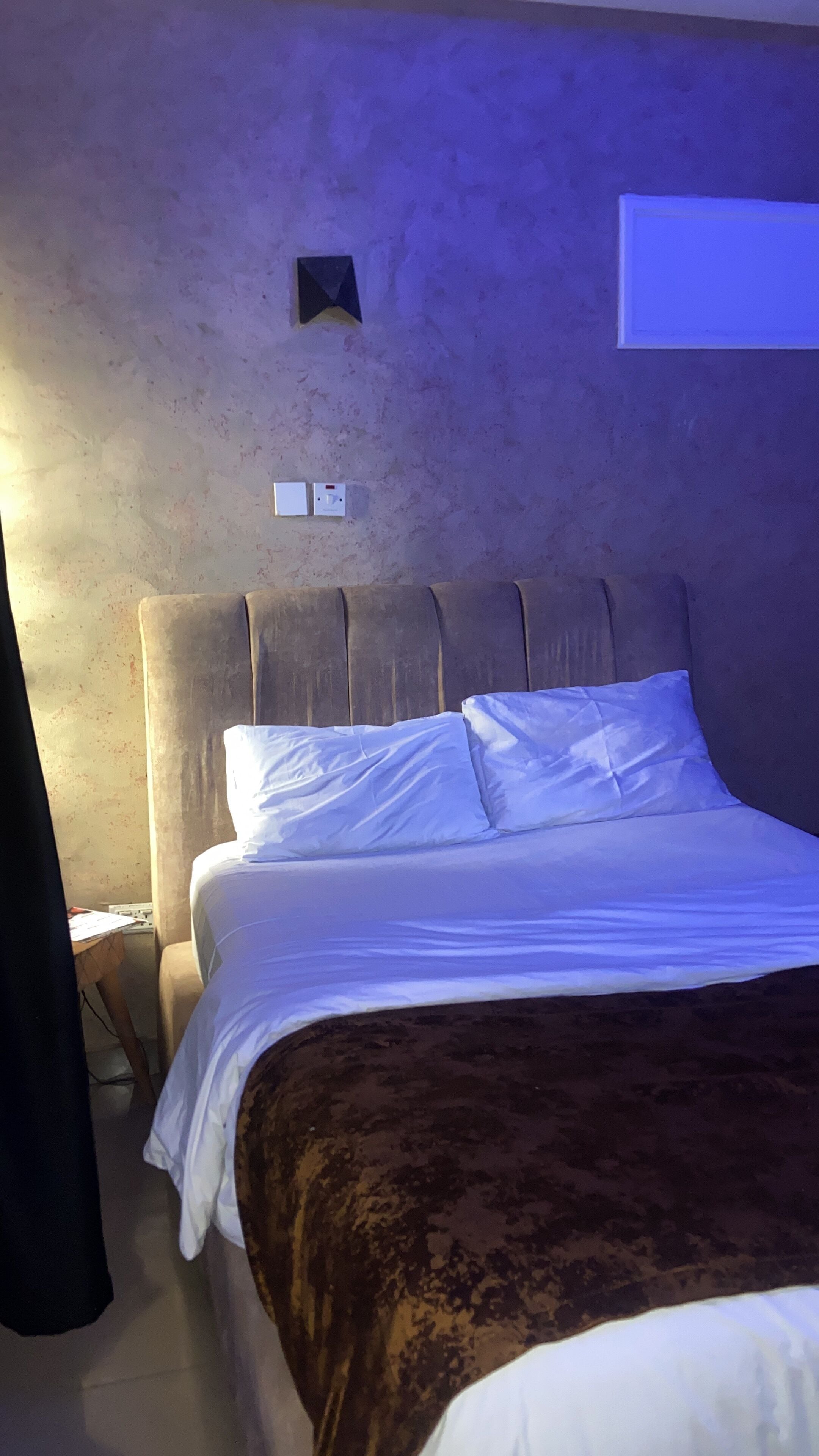 A bedroom with a bed, a headboard, a wall socket, and a window.