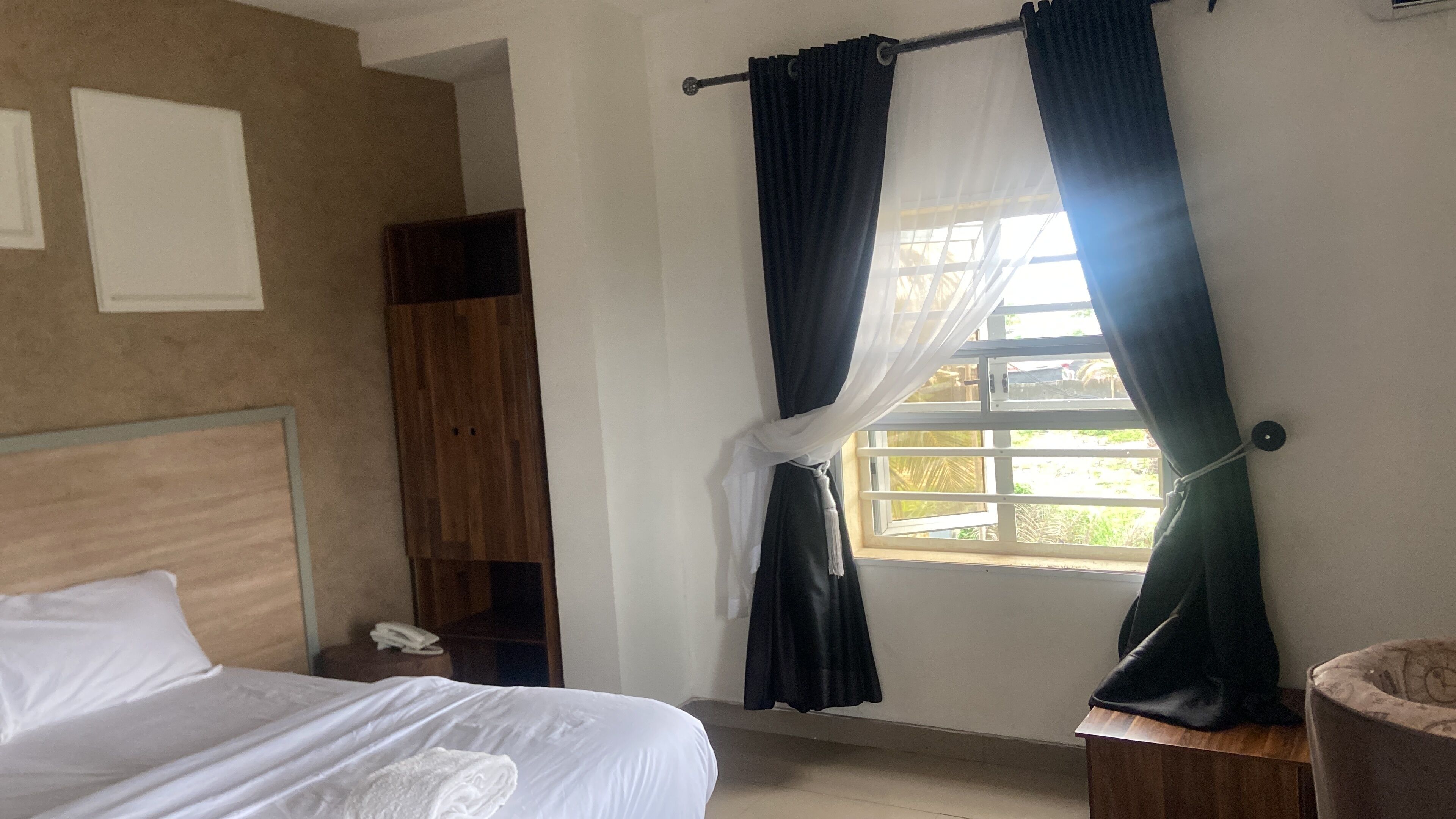 A hotel room with a bed, a wooden wardrobe, a window with curtains, and a small bedside table with a phone.