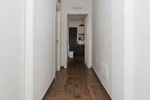 Un couloir au plancher en bois mène à une cuisine équipée de meubles de rangement et d’un luminaire suspendu.