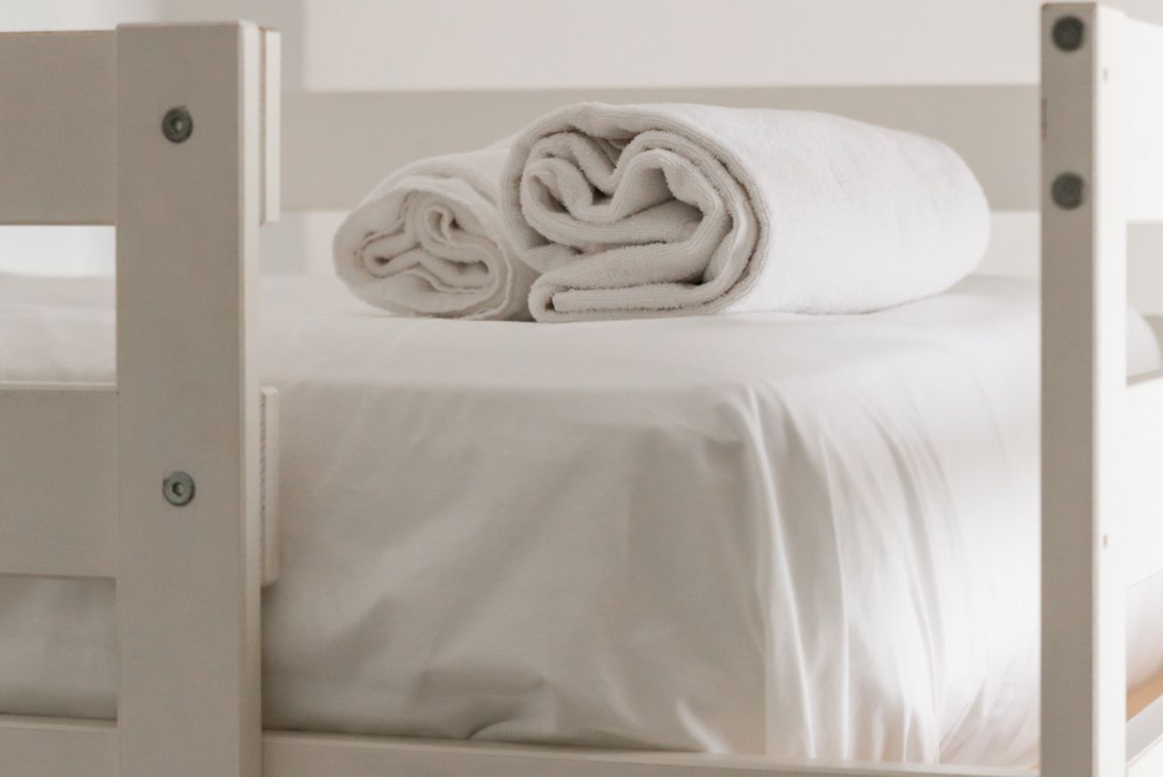 A white bed with a neatly rolled white towel on top.