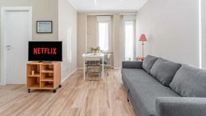 Ein modernes Wohnzimmer mit einem grauen Sofa, einem Fernseher auf einem Holzständer, einem Esstisch mit Stühlen und einer roten Lampe.