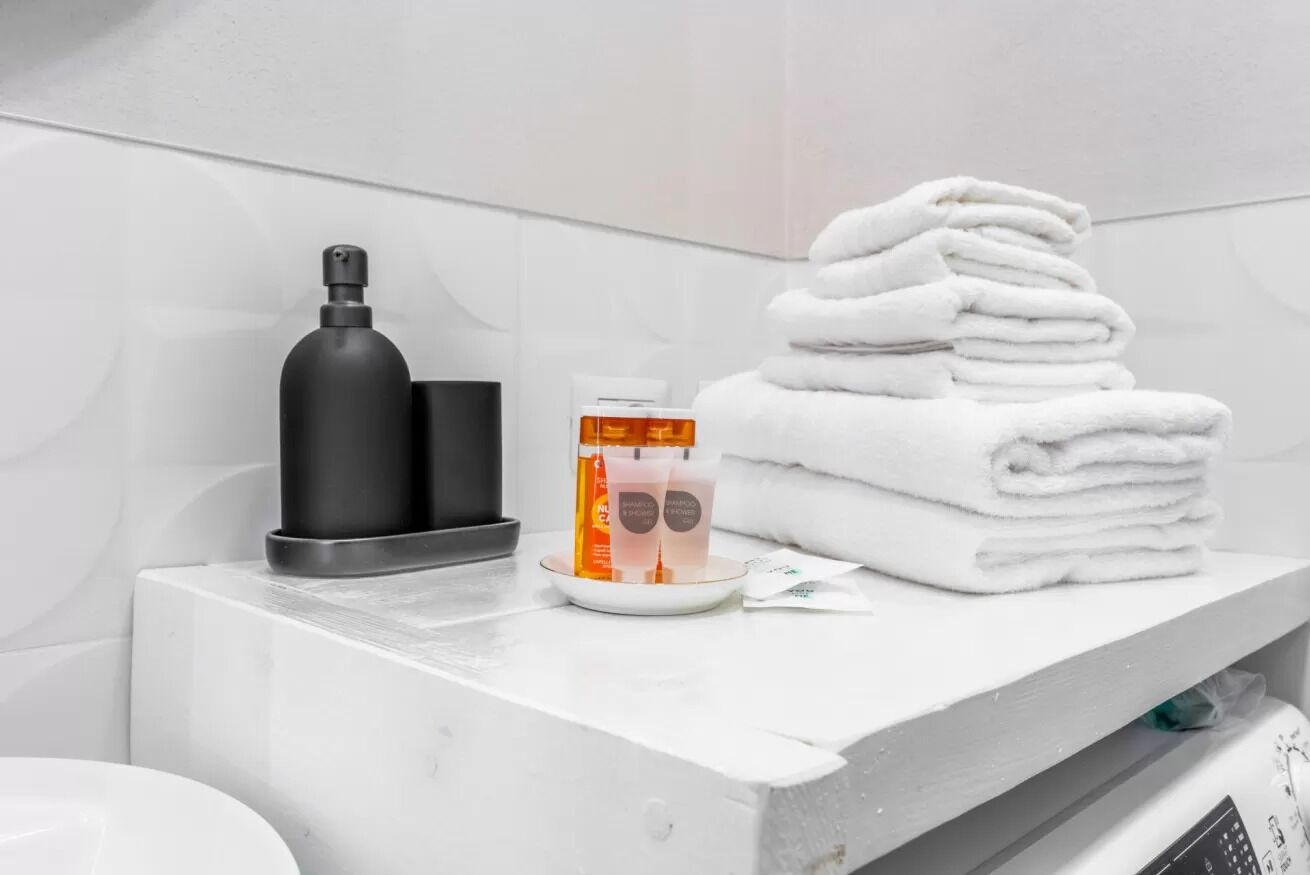 Une salle de bain avec un lavabo blanc, un distributeur de savon noir, une pile de serviettes blanches et une bouteille de shampoing.