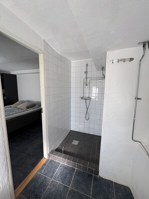 Ein kleines Badezimmer mit Fliesen, einer Dusche und einer Tür, sowie ein sichtbares Bett im angrenzenden Zimmer.