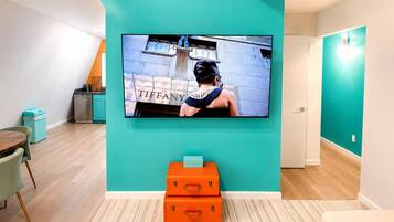 Un interior moderno con un televisor de pantalla plana montado en una pared verde azulada, una mesa de madera con sillas y un par de maletas naranjas en el suelo.