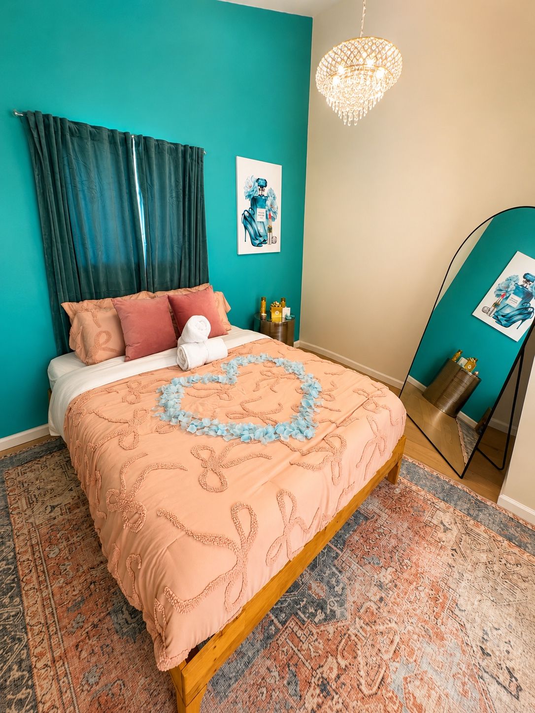 A bedroom with a teal wall, a bed with a peach-colored bedspread, a chandelier, and a mirror with a teal frame.
