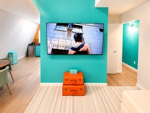 Um interior moderno com uma TV de tela plana presa a uma parede verde-azulado, uma mesa de madeira com cadeiras e um par de malas laranjas no chão.