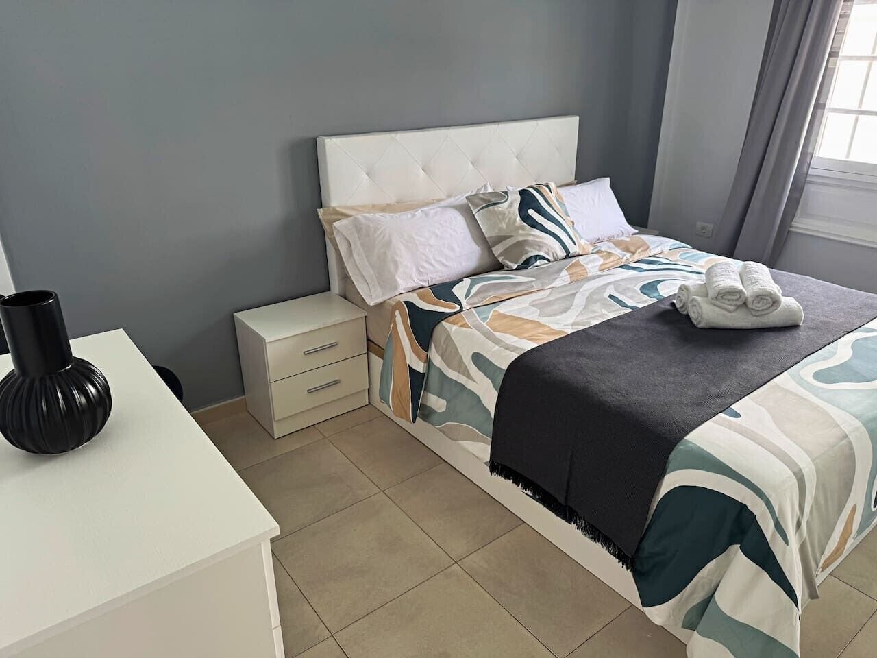 A bedroom with a bed, a bedside table, a drawer unit, and a window with curtains.