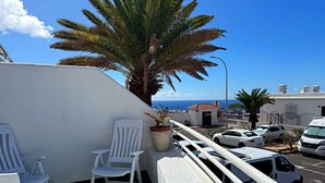A balcony with white chairs, a potted plant, and a view of the sea and buildings. - Apartment in Los Cristianos - Ocean View (Arona)