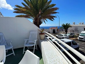 Ein Balkon mit weißen Stühlen, einem Blumentopf und Blick auf das Meer und Gebäude.