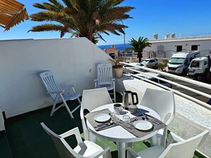 A rooftop terrace with a table set for two, white chairs, a potted plant, and a view of the sea and buildings. - Apartment in Los Cristianos - Ocean View (Arona)