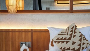 Una cama con ropa blanca, cabecera de madera y mesita de noche con un florero blanco.