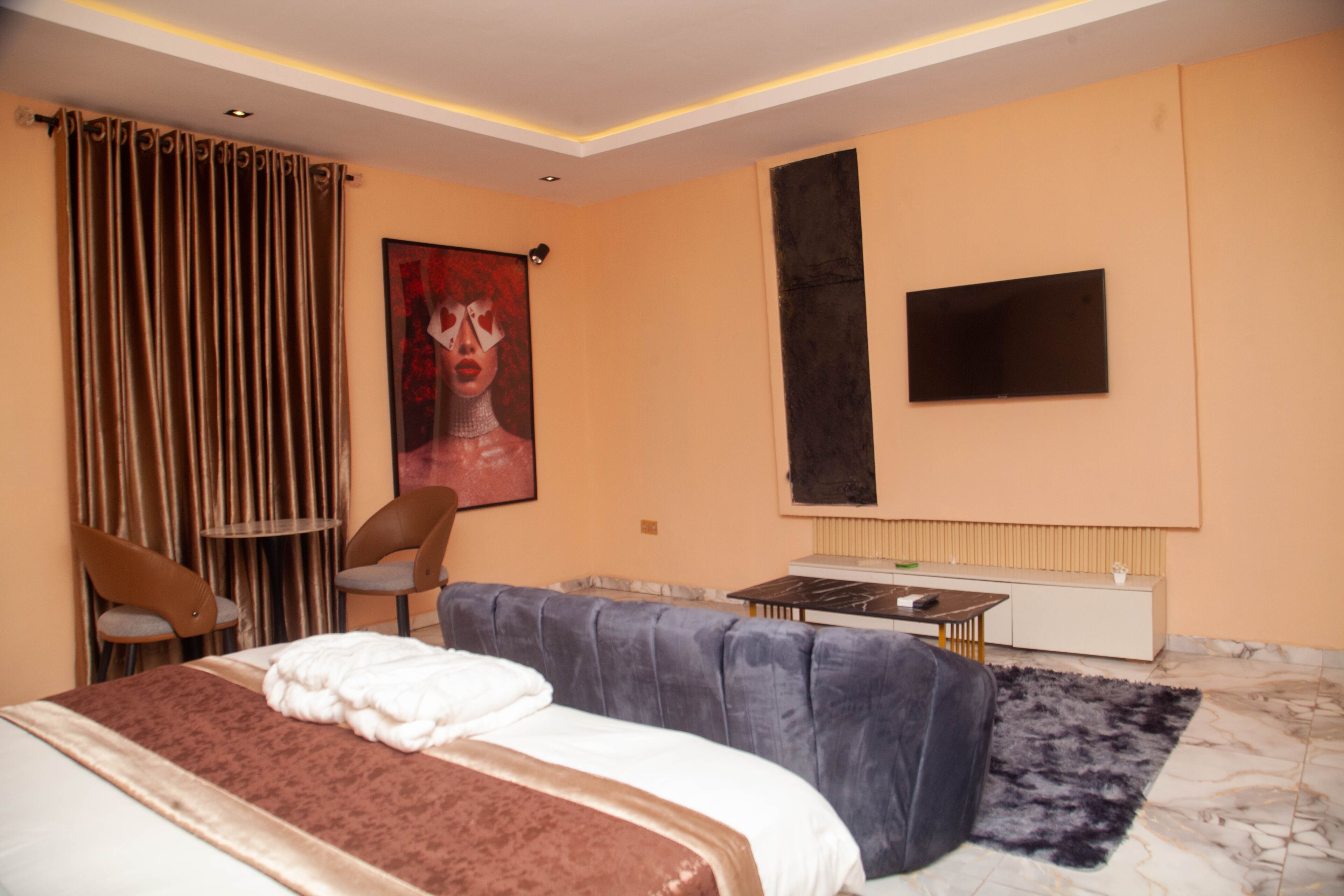Quarto de hotel com cama, sofá, mesinha, duas cadeiras, um quadro na parede e televisão fixada na parede.