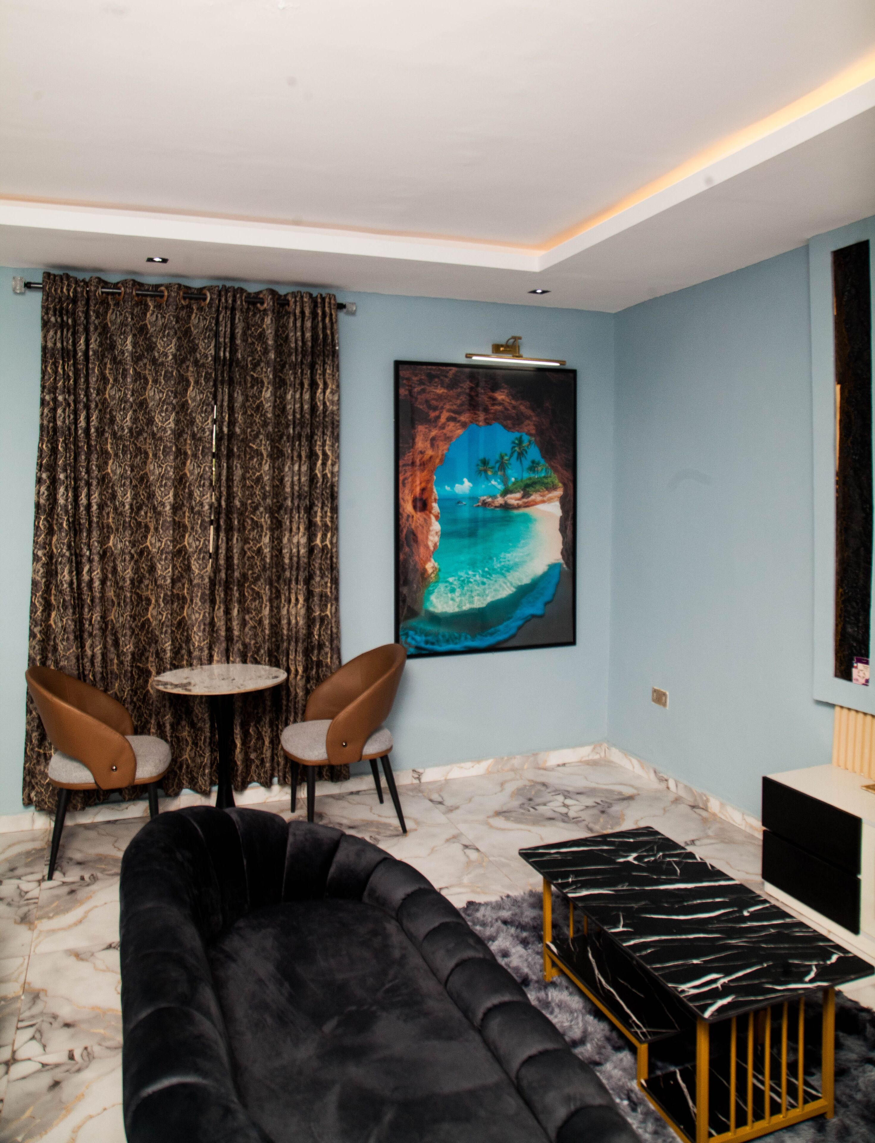 A living room with a black sofa, two brown chairs, a small round table, a large framed picture of a coastal scene, and a marble floor.
