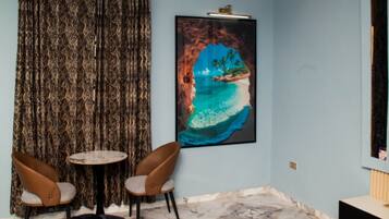 A living room with a black sofa, two brown chairs, a small round table, a large framed picture of a coastal scene, and a marble floor.