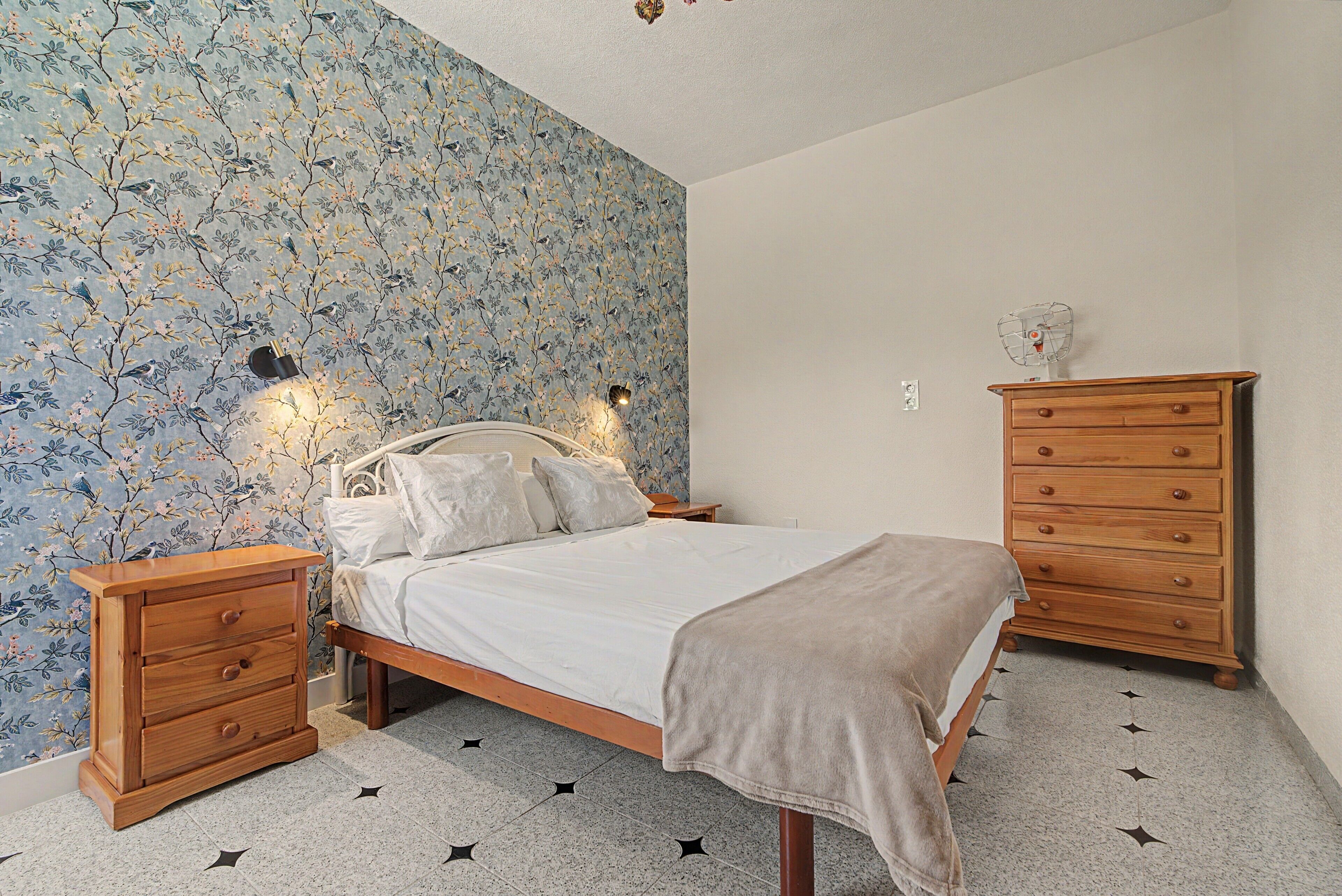 Un dormitorio con una cama, mesitas de noche de madera, un aparador y una pared con estampado floral.