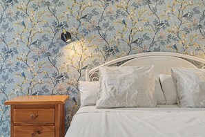 Une chambre avec un papier peint à motifs floraux, une table de chevet avec une lampe et un lit avec des oreillers.