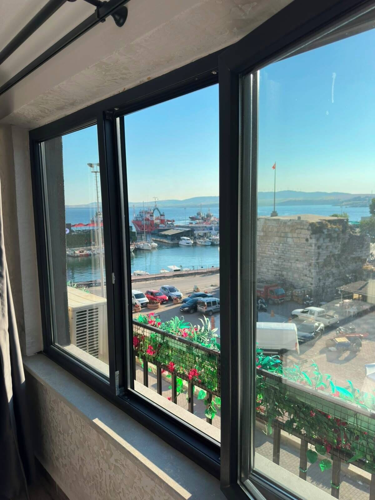 Vista desde una ventana con balcón, con vista a un puerto deportivo con barcos y una costa lejana.