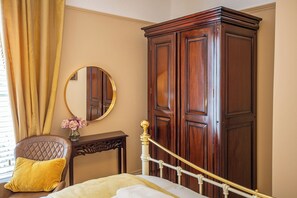 Un dormitorio con cama, un armario de madera, un espejo redondo, una mesita con un florero y una silla con cojín.