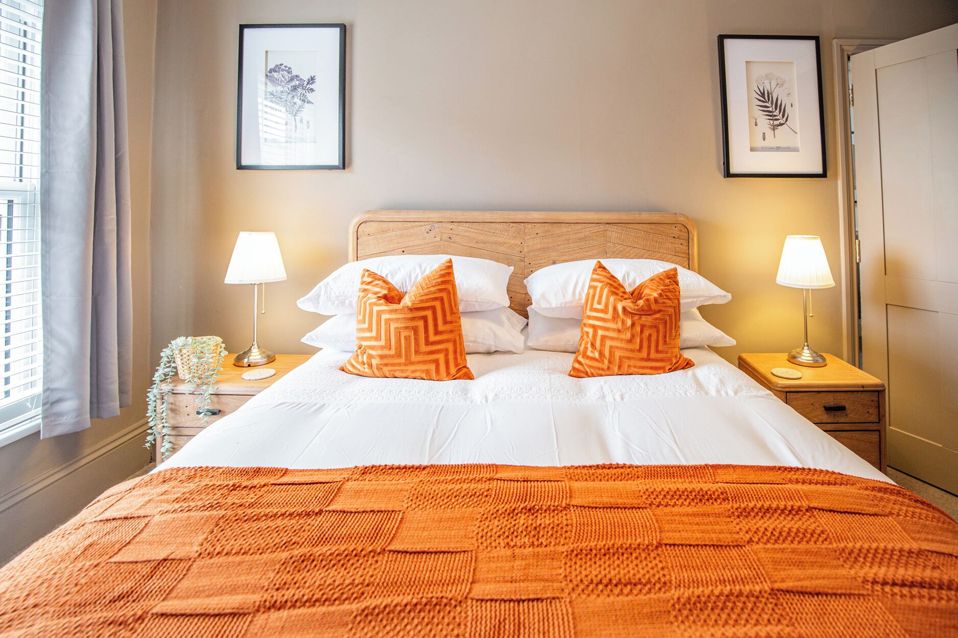 Una cama bien hecha con ropa blanca y almohadas estampadas en naranja, dos lámparas de noche e impresiones botánicas enmarcadas en la pared.