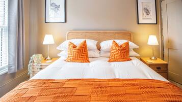 Una cama bien hecha con ropa blanca y almohadas estampadas en naranja, dos lámparas de noche e impresiones botánicas enmarcadas en la pared.