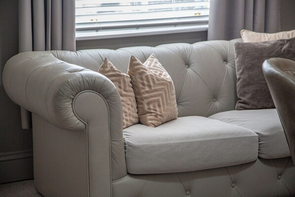 A light-colored tufted sofa with three patterned cushions, positioned near a window with blinds. - Hudson House (Durham)
