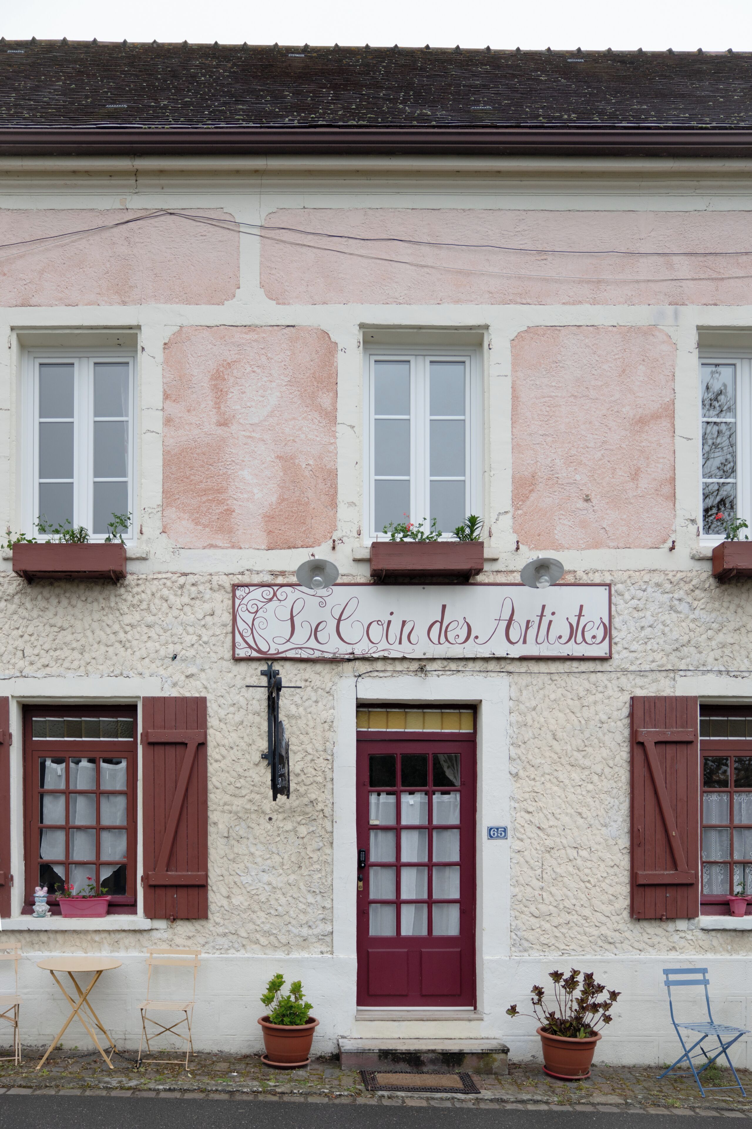 Ein Gebäude mit einem Schild, auf dem 'Le Coin des Artistes' steht, mit einer roten Tür, weißen Wänden und Topfpflanzen.