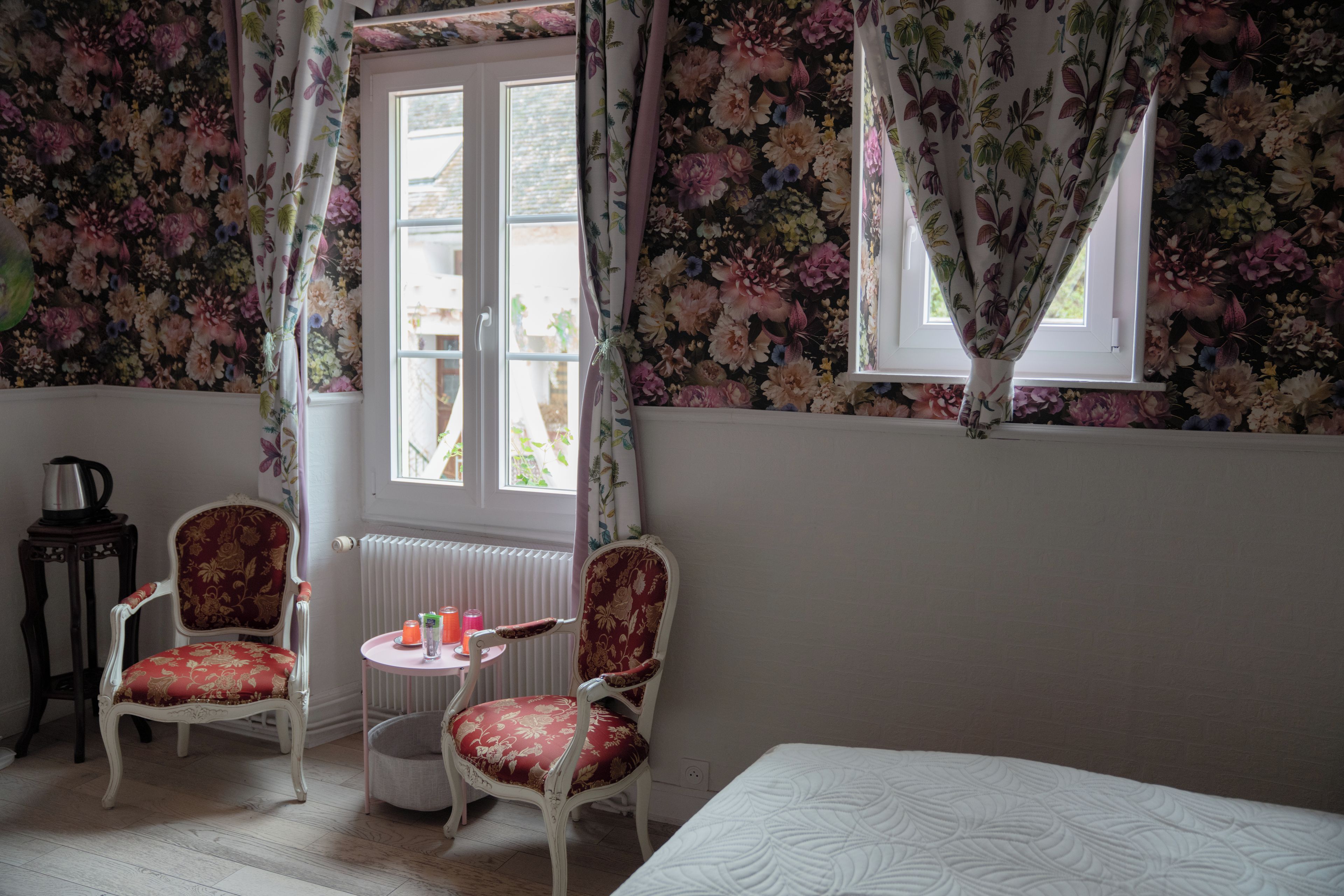 Een kamer met bloemenbehang, een bed, twee stoelen, een tafeltje met een waterkoker en een raam met gordijnen.