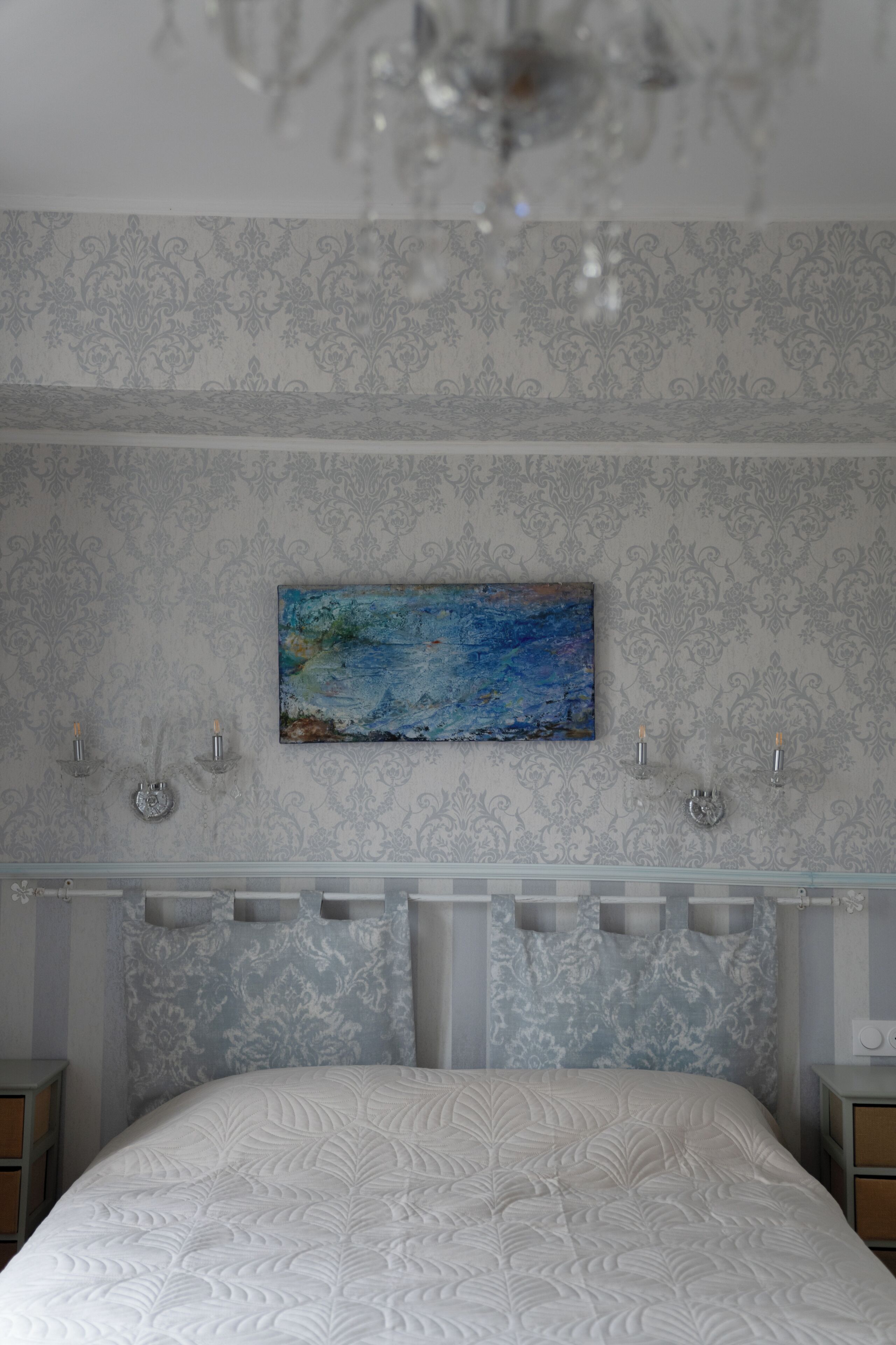 A bedroom with a bed, a painting on the wall, and a chandelier.
