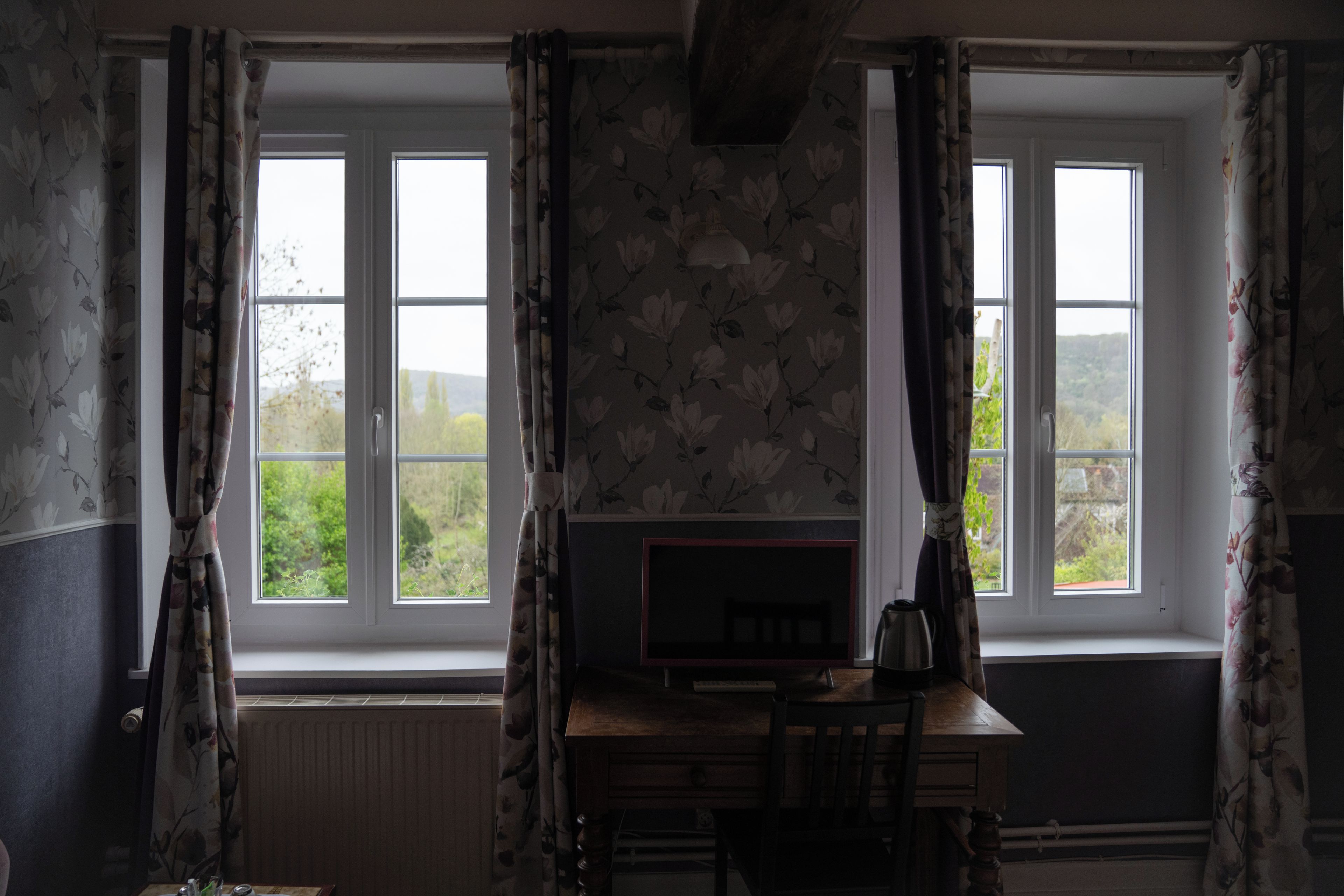 Ein Zimmer mit zwei Fenstern, einem Heizkörper, einem Holztisch mit Stuhl und einem Fernseher.
