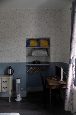 A room with a bed, a bedside table with a kettle, a heater, a desk with a computer, and a shelf with folded blankets.