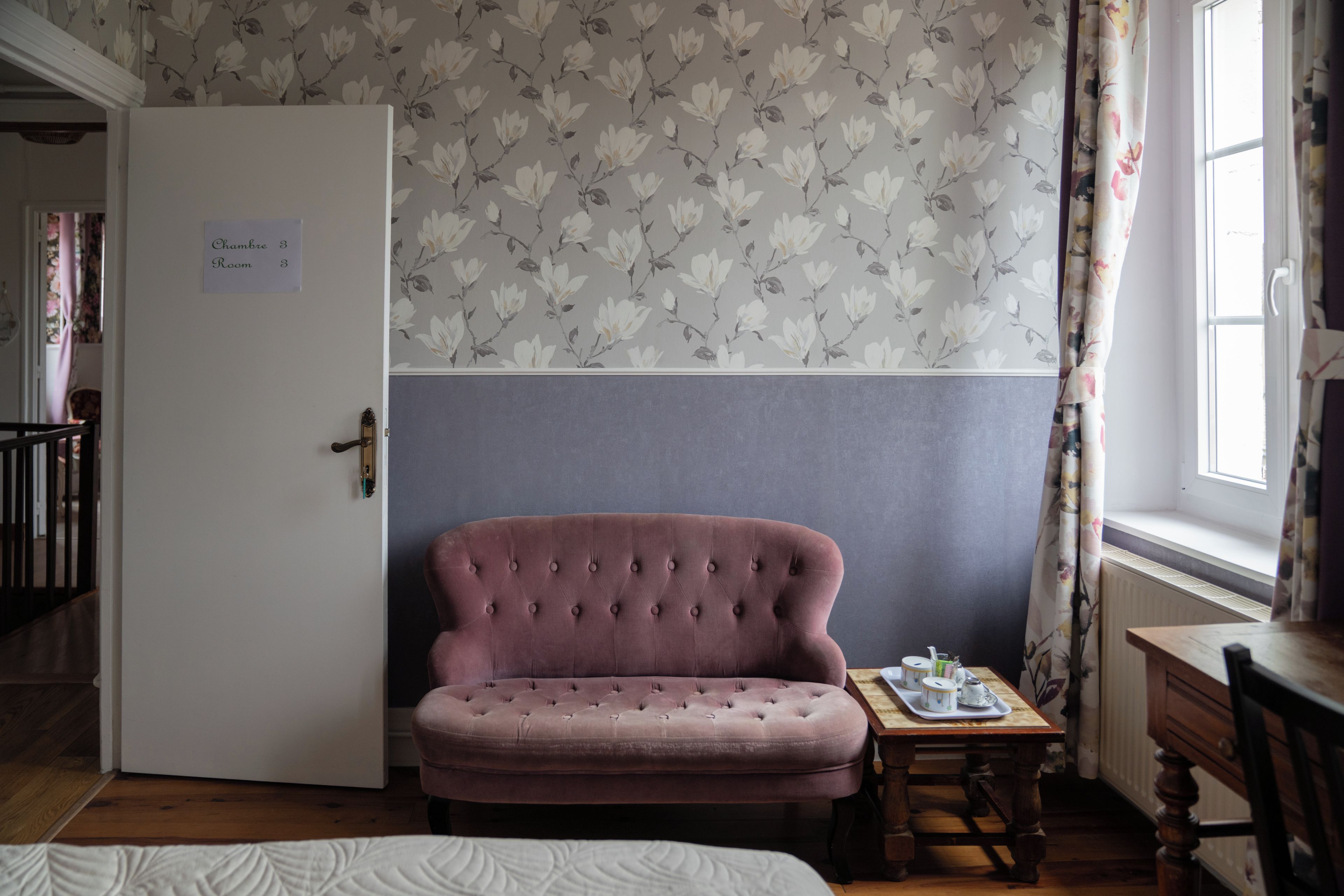 Une pièce avec une porte fermée, un fauteuil rose recouvert de tissu, une petite table avec un plateau, et une fenêtre avec des rideaux.