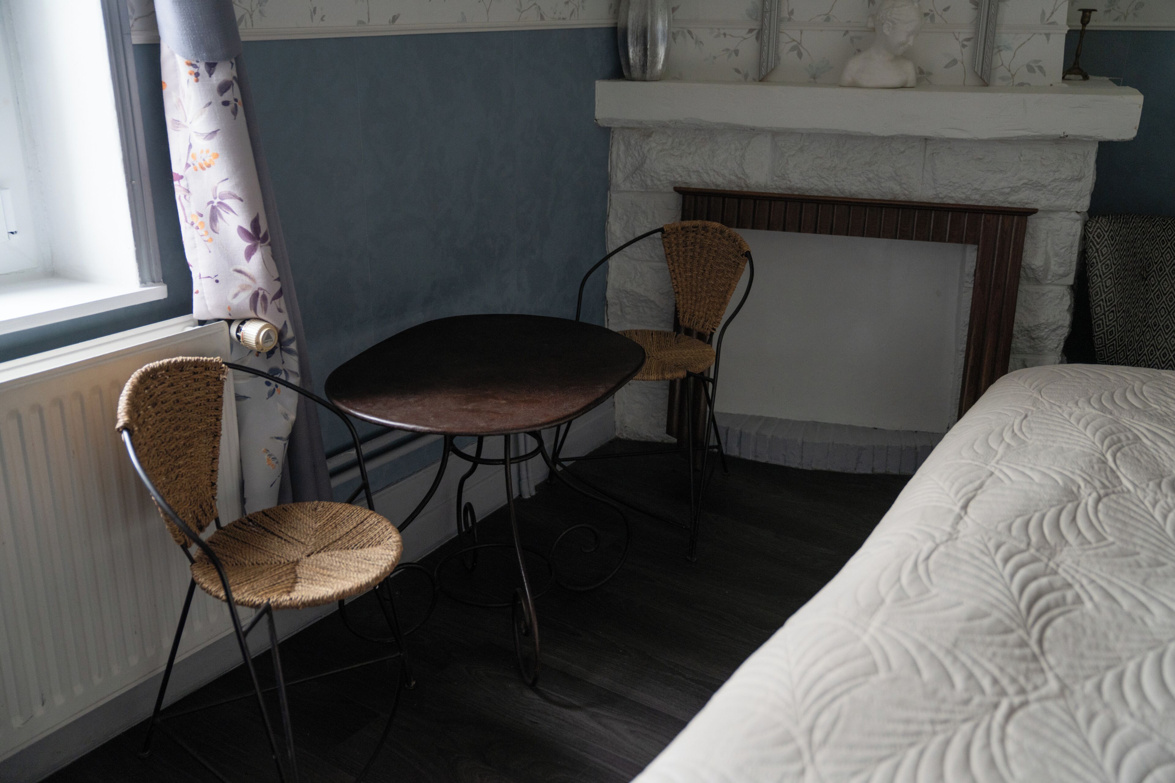Een kamer met een bed, een open haard, een tafel en twee rieten stoelen.