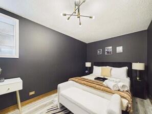 Ein modernes Schlafzimmer mit dunkler Wand, einem weißen Bett, zwei Nachttischlampen, einer Deckenleuchte, einem Schreibtisch und einem Teppich mit ZEBRA-MUSTER.
