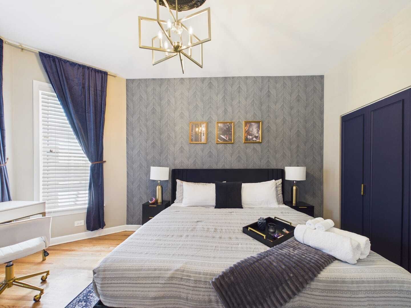 Quarto de hotel com cama, mesas de cabeceira, escrivaninha, cadeira, guarda-roupa e um candelabro.