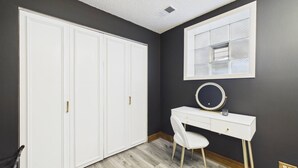 Quarto com um guarda-roupa branco grande, uma escrivaninha com cadeira, um espelho redondo e uma janela com mosquiteiro.
