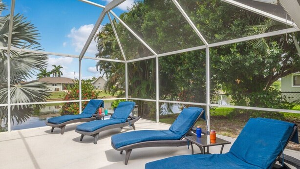A covered outdoor area with lounge chairs and a table, surrounded by a glass enclosure with a view of greenery and a house. - Coral Princess Waterfront Pool & Hot Tub (Cape Coral)