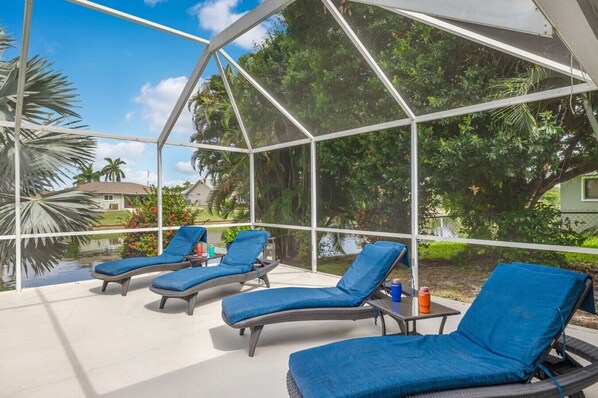 A covered outdoor area with lounge chairs and a table, surrounded by a glass enclosure with a view of greenery and a house.