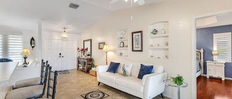 A spacious living area with a white sofa, a dining table, chairs, a ceiling fan, and a small side table with a lamp.