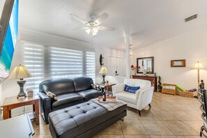 A living room with a black leather sofa, a white armchair, a ceiling fan, and a large window with blinds. - Coral Princess Waterfront Pool & Hot Tub (Cape Coral)